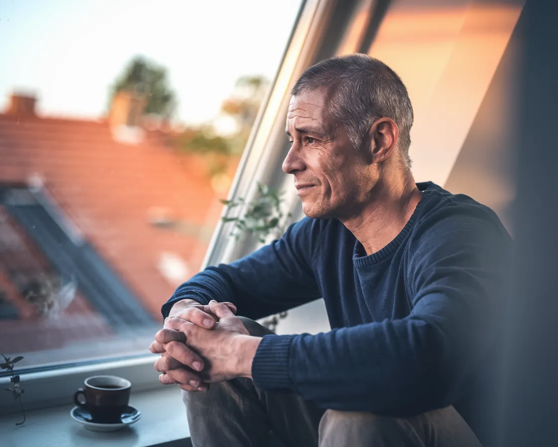 Imagem gerada por IA: homem na janela de telhado com chávena de café, olhar pensativo — Wohnperspektive